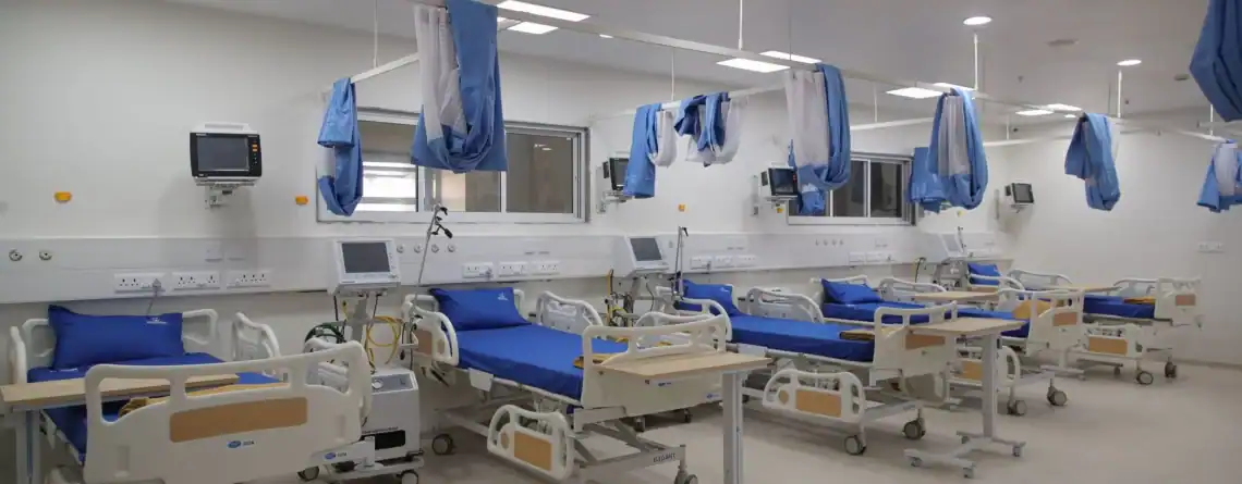 A brightly lit hospital recovery ward with multiple modern patient beds arranged in a row, each equipped with blue mattresses, bedside monitors, ventilator units, and movable overbed tables. Blue privacy curtains hang from ceiling-mounted tracks, and the room features clean white walls, medical outlets, and large windows providing visibility from the nursing area.