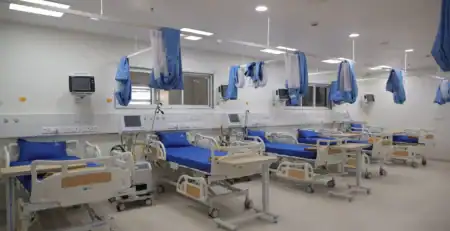 A brightly lit hospital recovery ward with multiple modern patient beds arranged in a row, each equipped with blue mattresses, bedside monitors, ventilator units, and movable overbed tables. Blue privacy curtains hang from ceiling-mounted tracks, and the room features clean white walls, medical outlets, and large windows providing visibility from the nursing area.