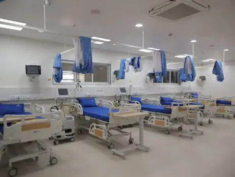 A brightly lit hospital recovery ward with multiple modern patient beds arranged in a row, each equipped with blue mattresses, bedside monitors, ventilator units, and movable overbed tables. Blue privacy curtains hang from ceiling-mounted tracks, and the room features clean white walls, medical outlets, and large windows providing visibility from the nursing area.